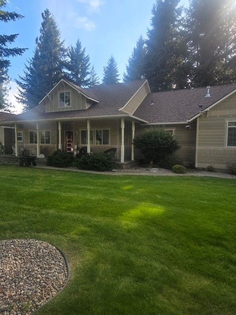 Suburban house with a green lawn, covered porch, and tall pine trees in the background.