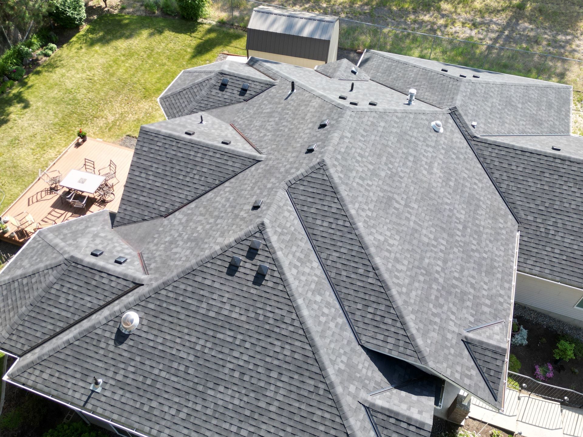 Aerial view of a gray shingle roof with multiple gables and skylights, beside a backyard patio and greenery