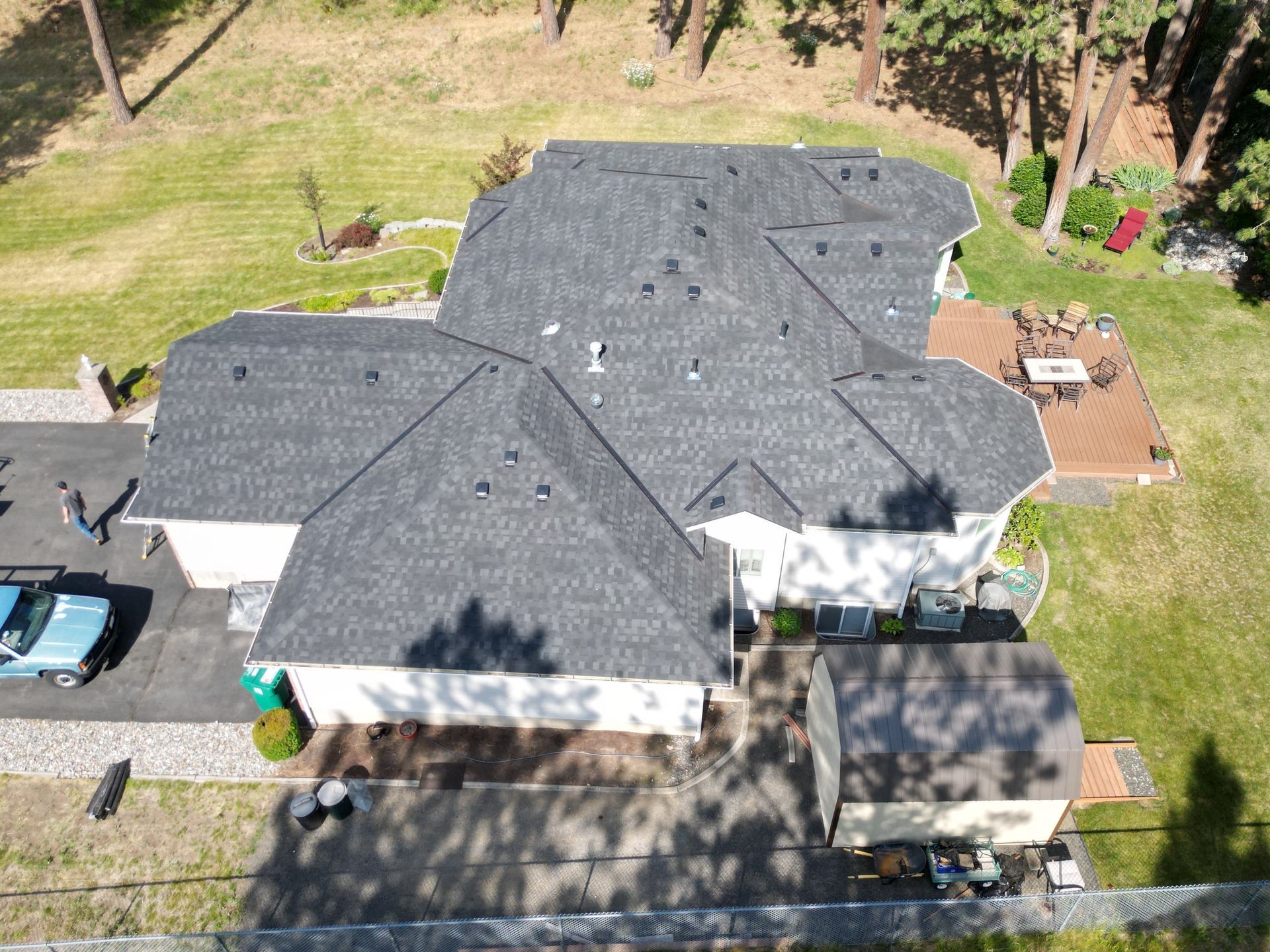 Aerial view of a house with dark gray roof, driveway, and surrounding yard with trees and shadows. Aerial view of a house with dark gray roof, driveway, and surrounding yard with trees and shadows.