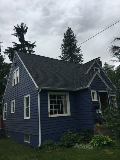 Blue two-story house with white trim and steep roof, seen from the side amid trees and greenery.