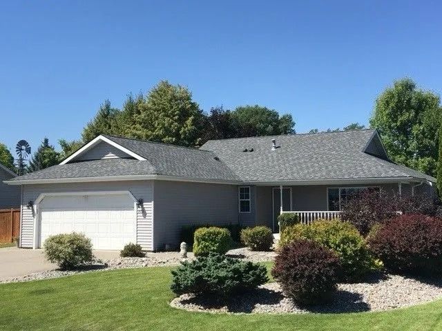 Suburban single-story house with a two-car garage, gray roof, and landscaped front yard under a blue sky