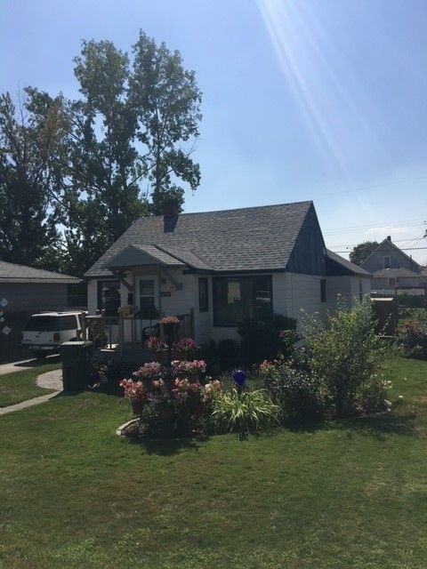 Small white house with a gray roof, front garden, and large trees in bright sunlight.