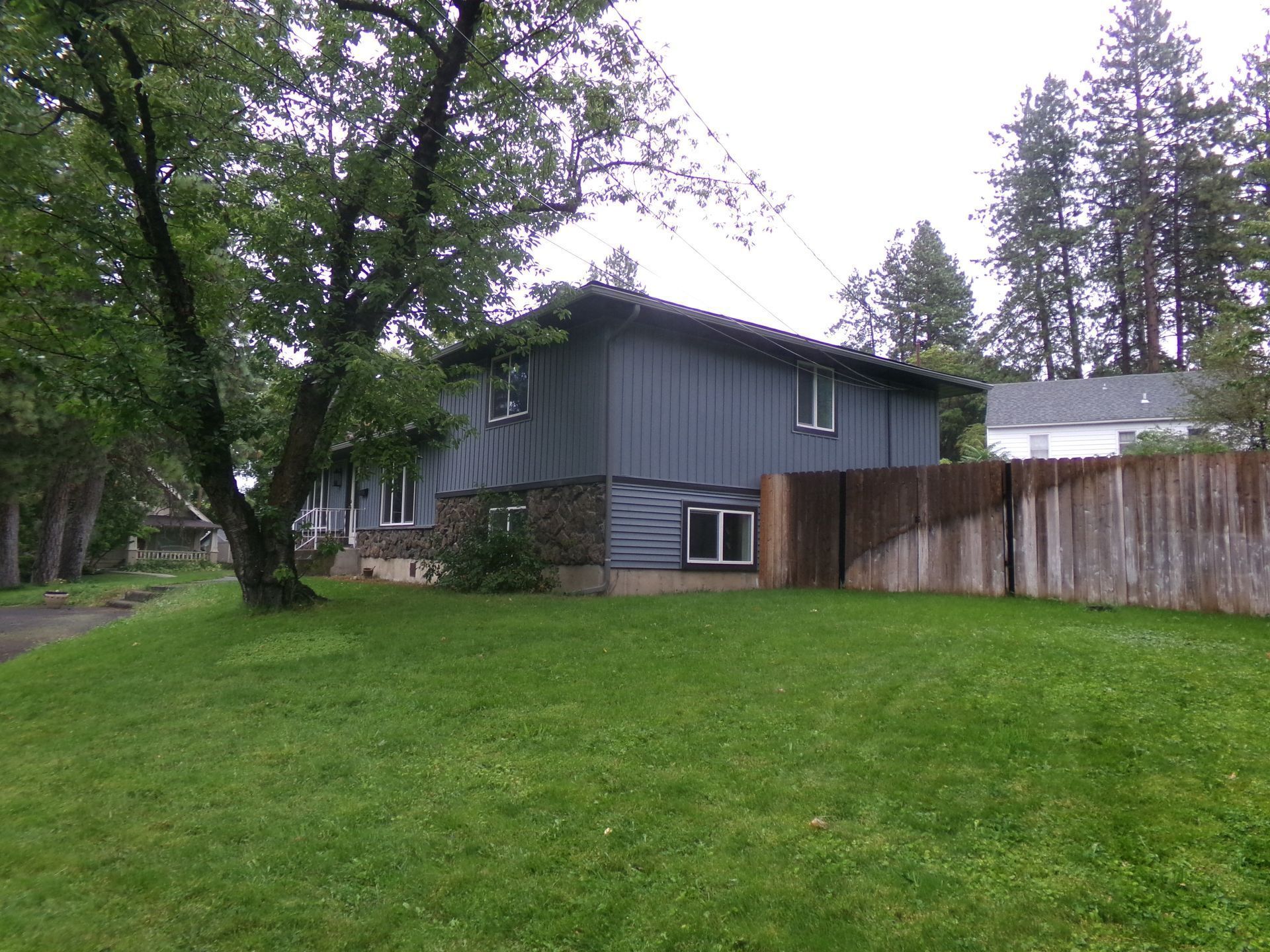 Gray house with a large green lawn, wooden fence, and trees in a residential yard