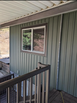 Covered porch with green siding, white-trimmed window, and black railing overlooking a rocky hillside.
