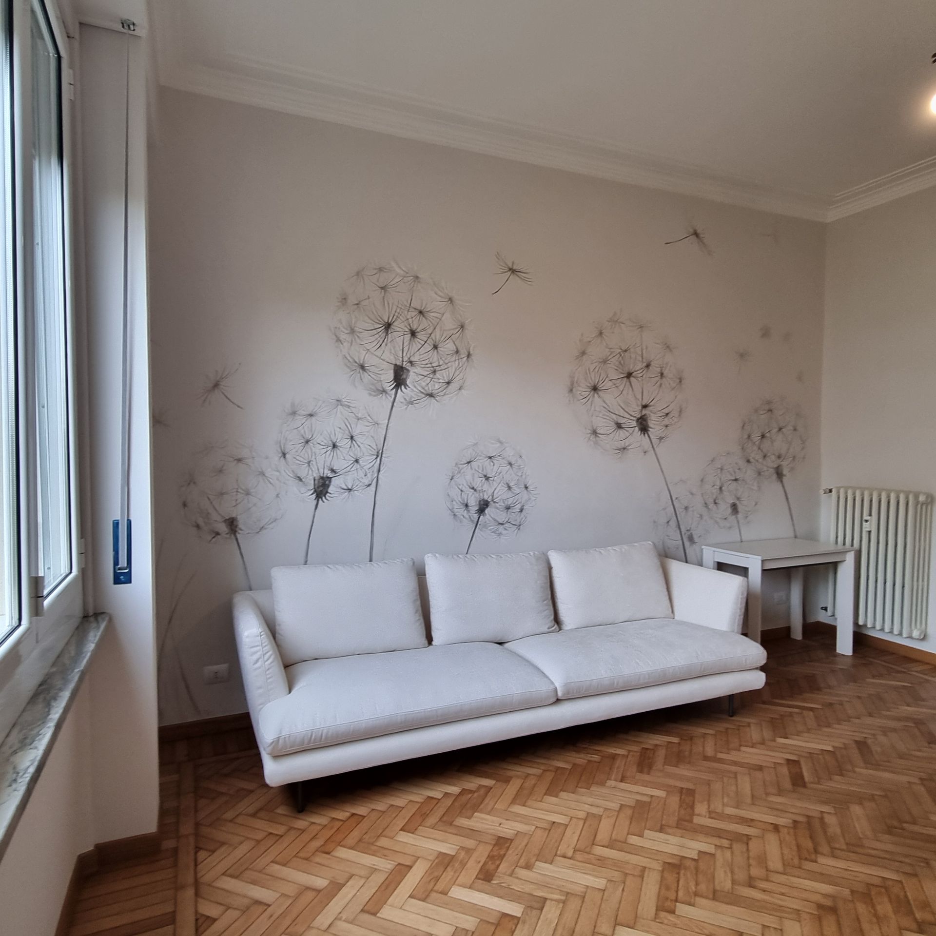 Luminoso soggiorno con divano bianco, adesivi murali a forma di dente di leone, pavimento in legno e ampia finestra.
