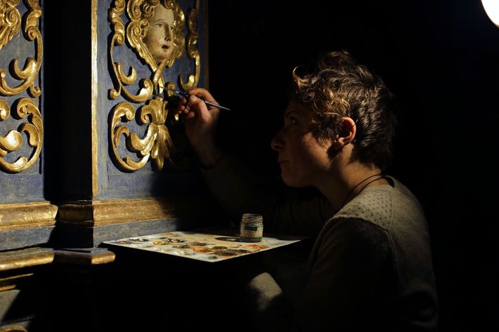 Artista che dipinge un pannello murale blu dorato in condizioni di scarsa illuminazione, con pennelli e tavolozza su un tavolo.