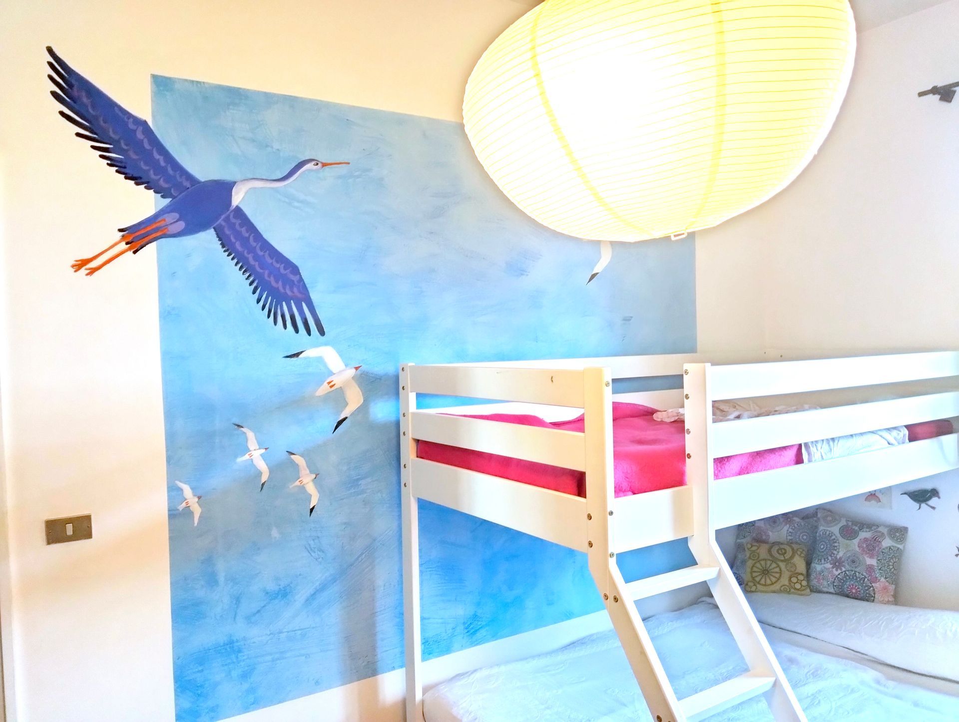 Camera da letto per bambini con letto a castello bianco, murale raffigurante un cielo azzurro e uccelli dipinti sulla parete.