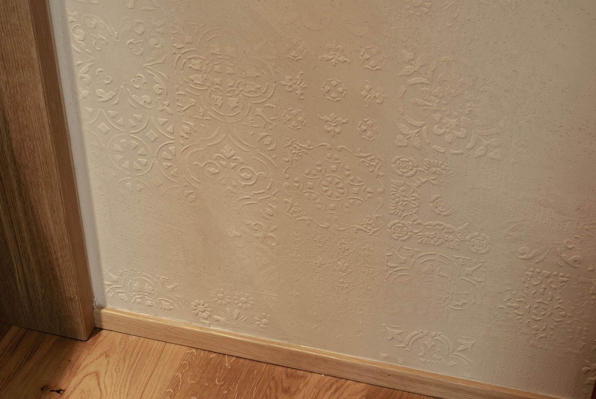 Angolo di una stanza con parete beige texturizzata, battiscopa in legno e pavimento in parquet.