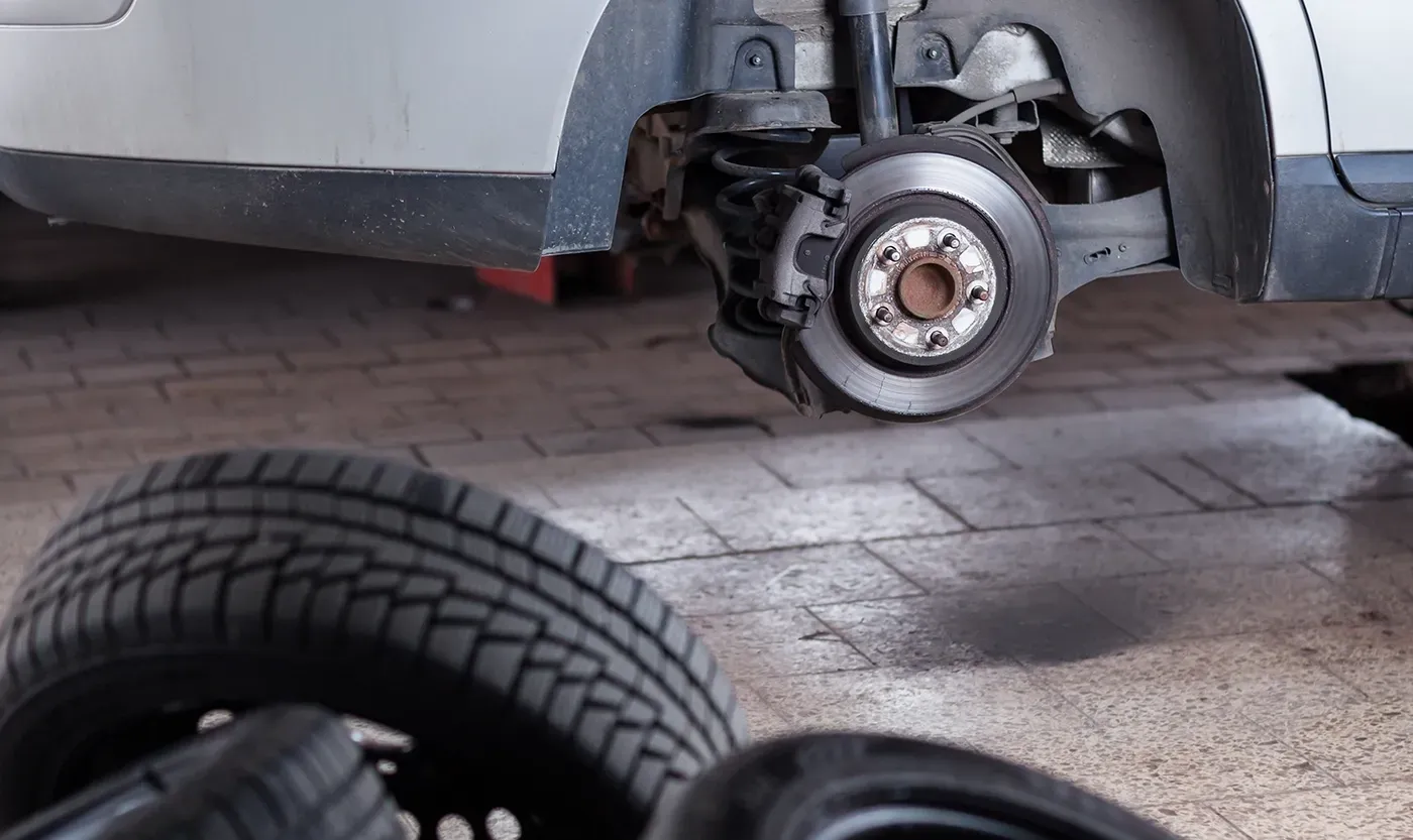 Car with a wheel removed, exposing the brake assembly in a garage; loose tire in foreground.