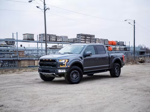 A gray Ford F-150 Raptor truck parked on a gravel lot in front of industrial warehouse storage racks.