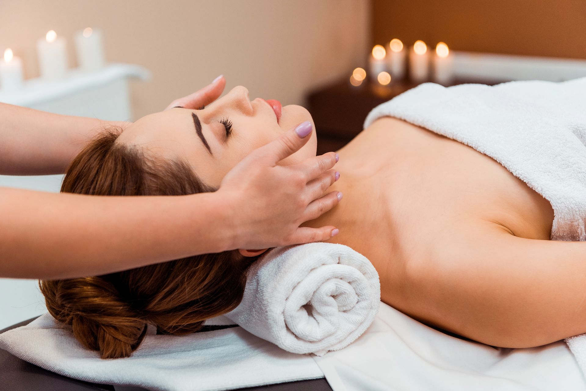 Una donna riceve un massaggio al viso su un lettino da spa, con asciugamani bianchi arrotolati e candele sullo sfondo.