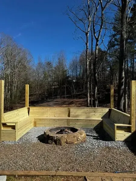Outdoor wooden seating surrounds a stone fire pit, gravel ground, trees in background, blue sky.