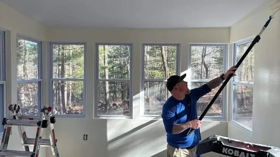 Person painting a wall in a room with large windows; using a paint roller attached to a telescoping pole.