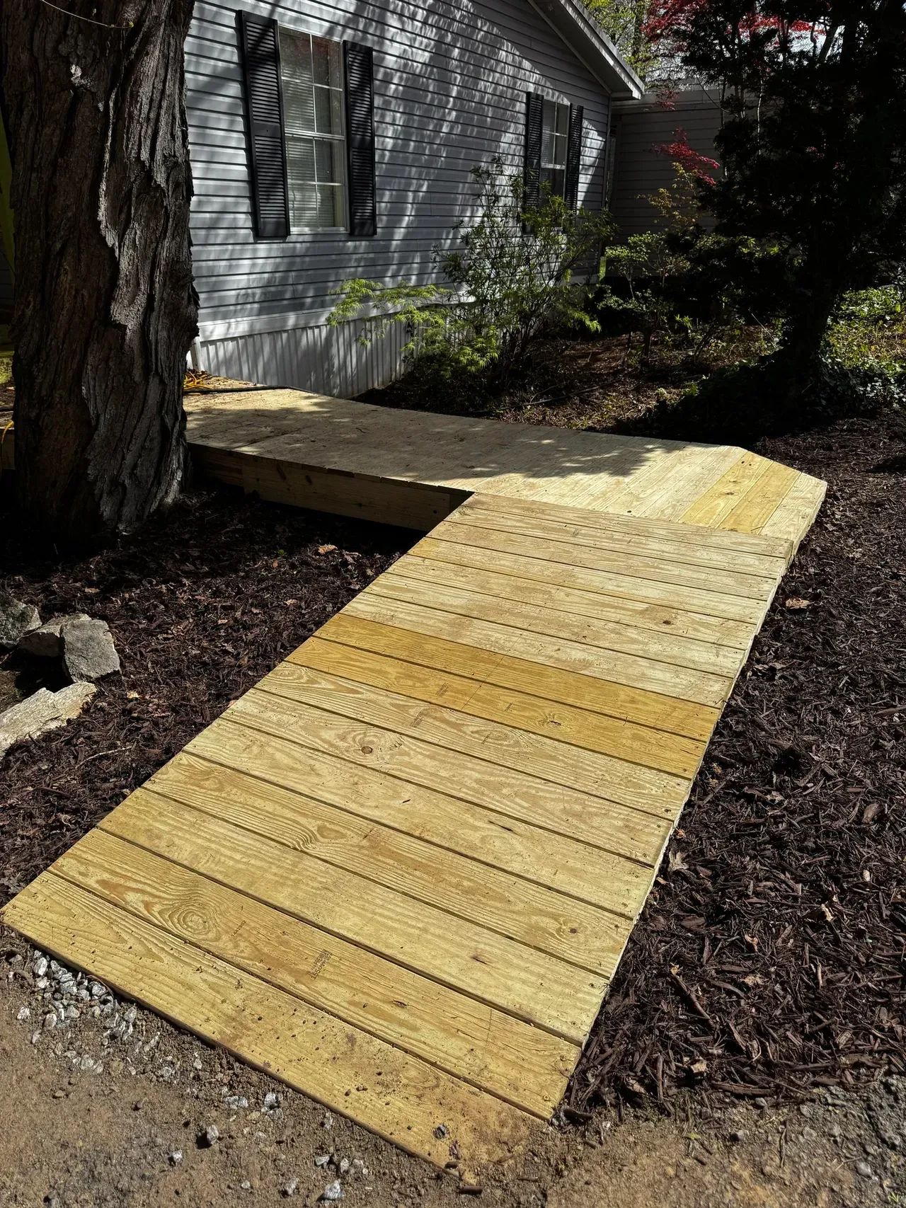 Wooden ramp leading to the entrance of a light gray house. Brown mulch and a tree are nearby.