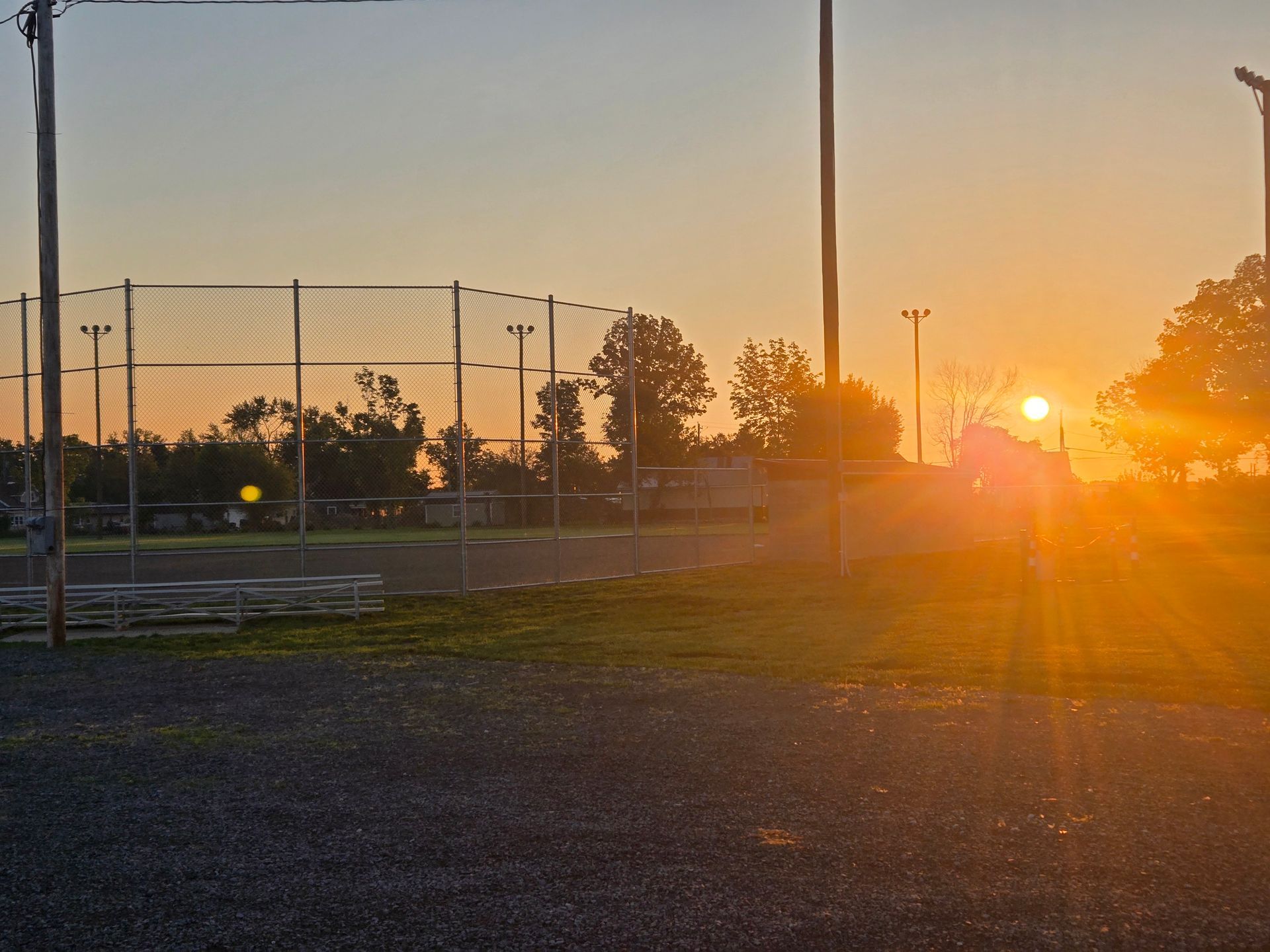 Sunset over a sports field with chain-link fence and trees silhouetted against orange sky