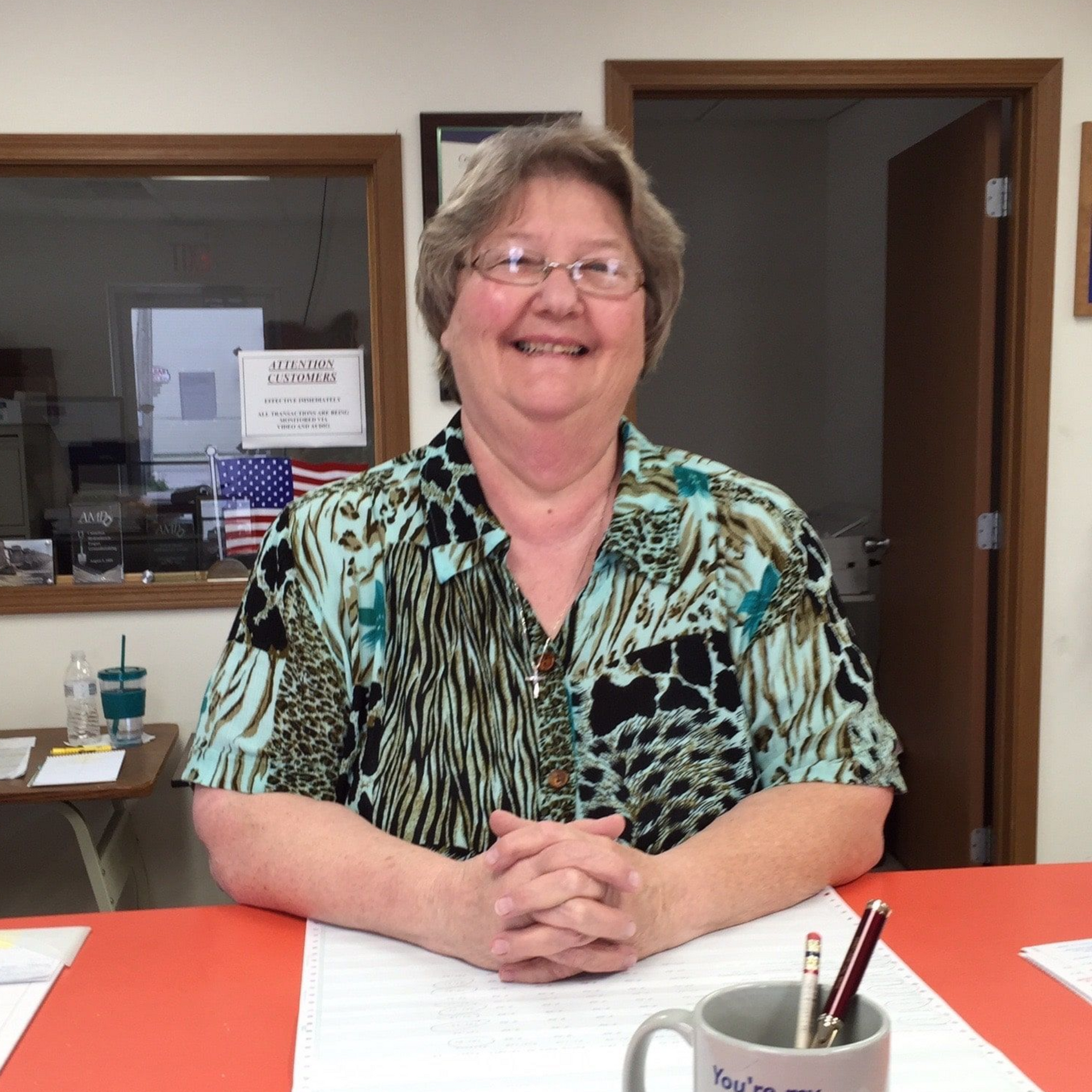 Smiling woman seated at a desk in an office, hands clasped, with papers and a pen holder in front.