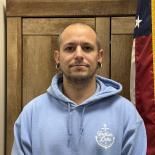 Man in light blue hoodie standing before wooden paneling with an American flag at right