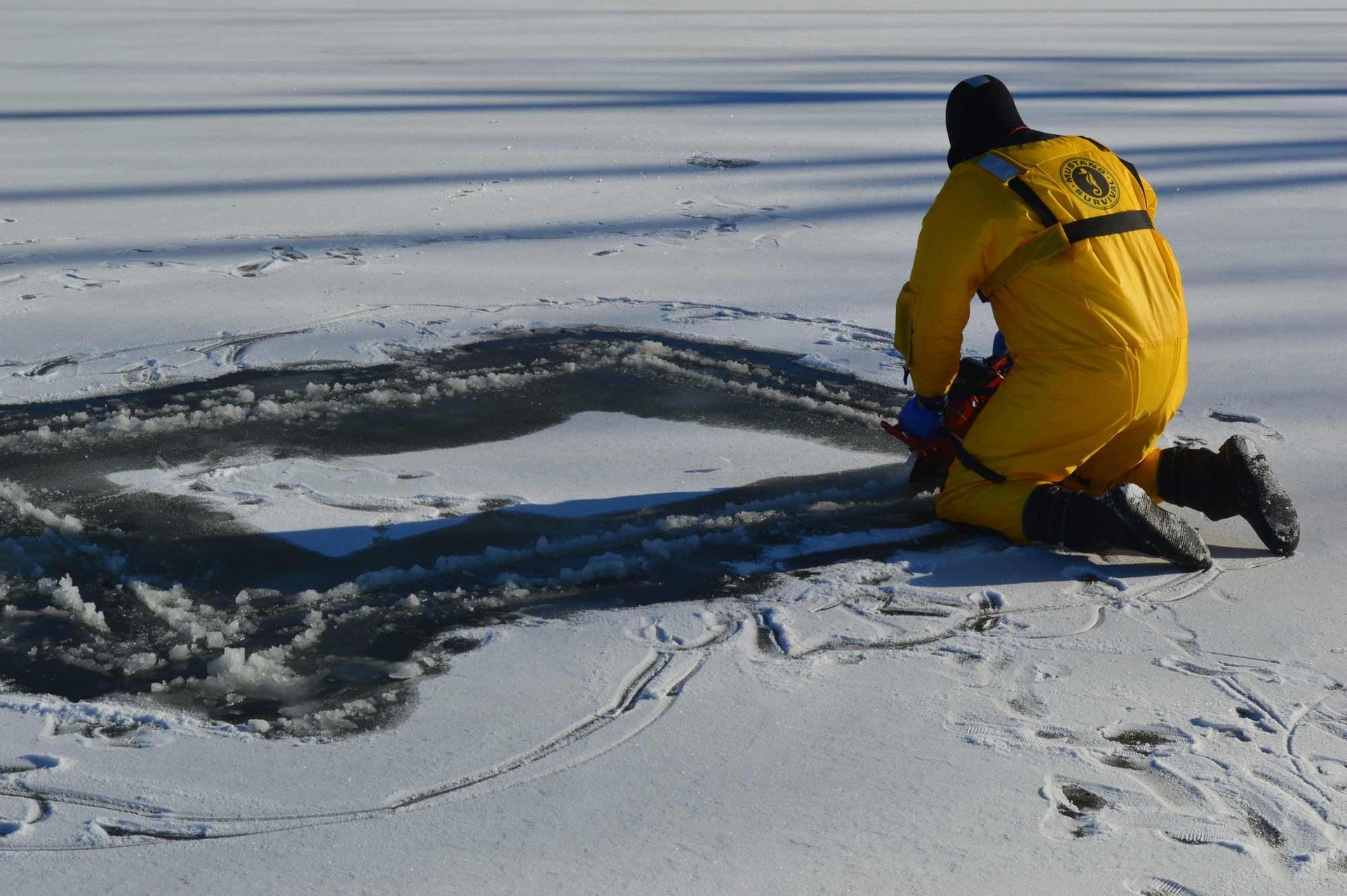 Person in yellow winter gear kneeling on ice beside an ice-fishing hole, snowy frozen lake around them