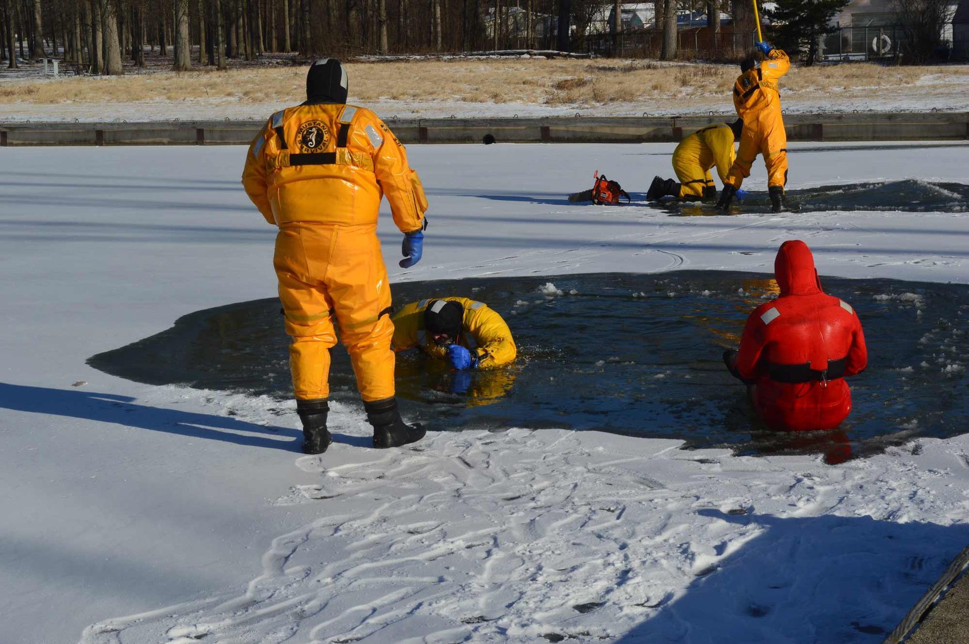 Workers in yellow and red cold-weather suits inspect a frozen lake near an open hole