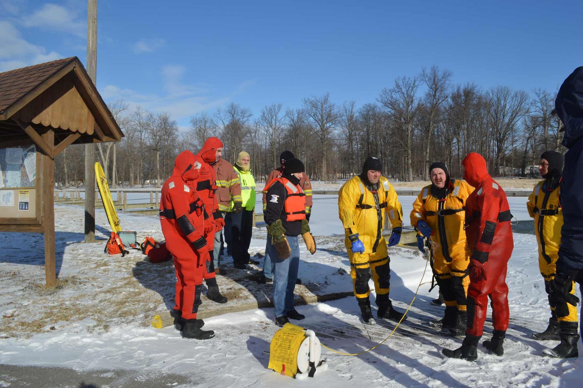 Workers in red and yellow rescue suits gather on a snowy field near a wooden shelter.