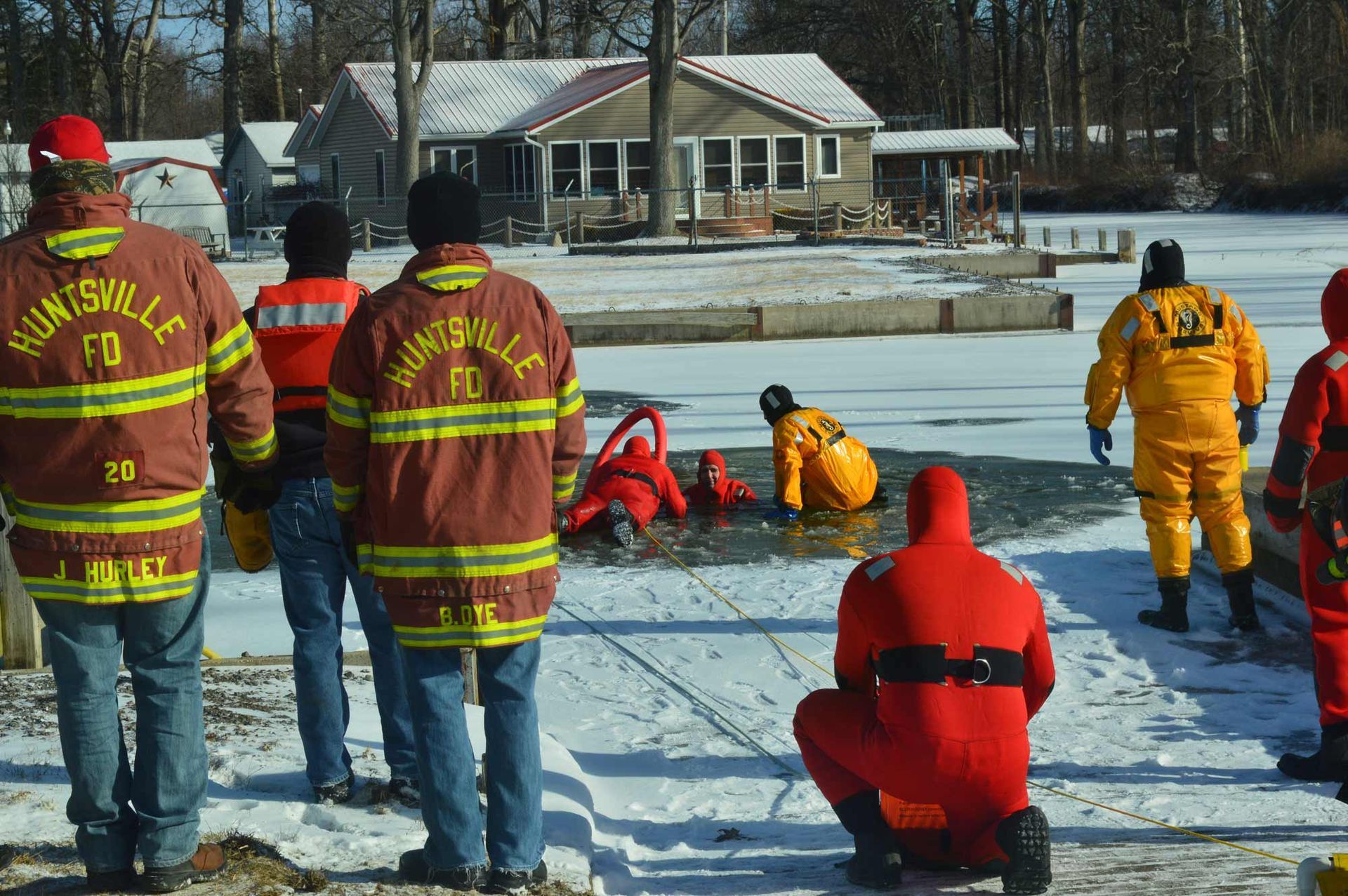 Rescue workers in bright winter gear assisting people on a frozen lake near houses and trees