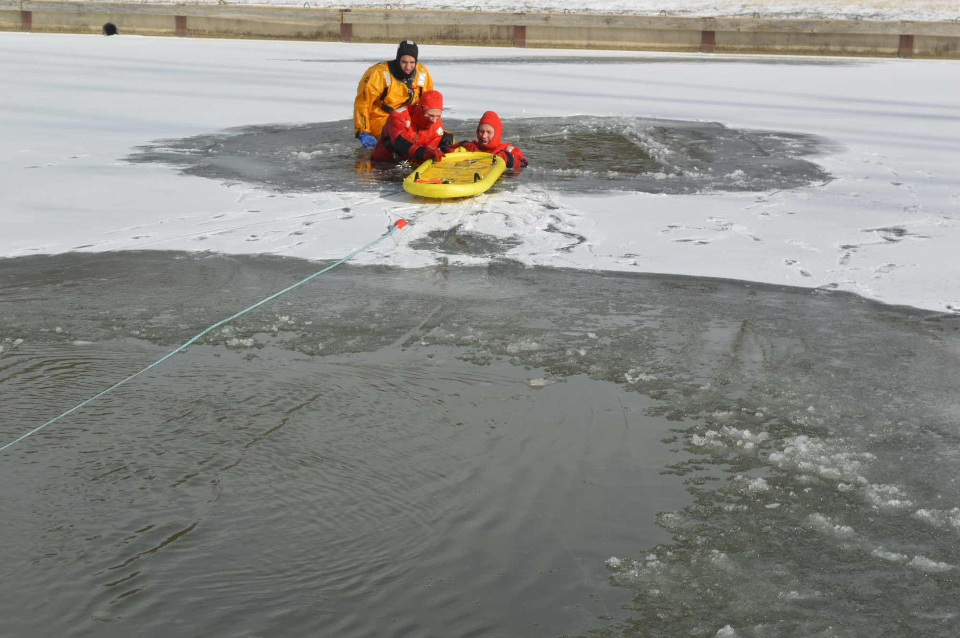 Rescue workers in orange gear on a frozen lake, using a yellow sled near an ice hole