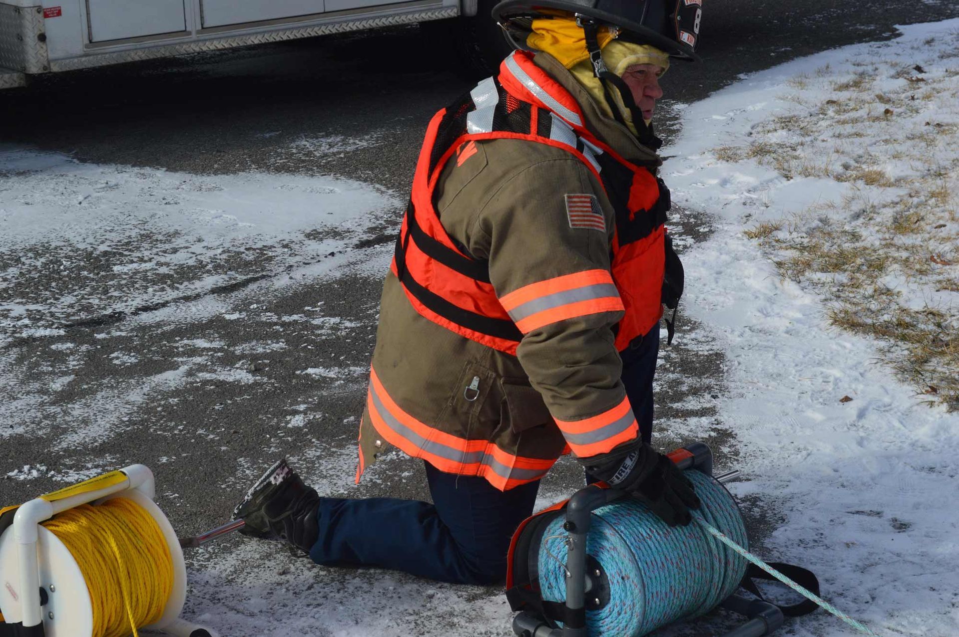 Firefighter kneeling on icy ground beside a blue hose reel near a truck