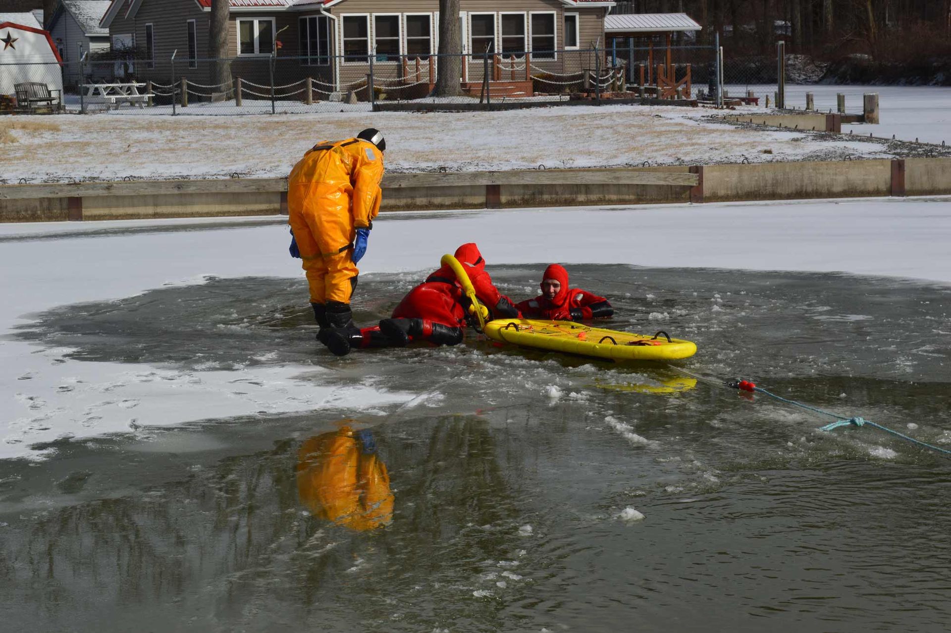 Two rescue workers in orange gear on ice beside a yellow rescue sled near water and buildings