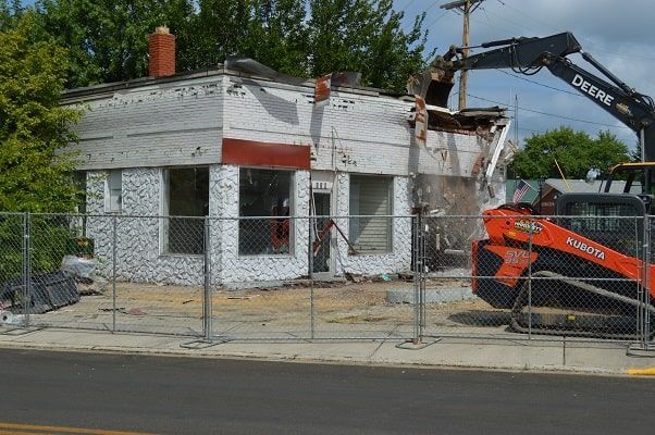 Demolished white house behind a chain-link fence, with an excavator tearing down the roof.