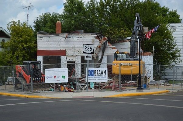 Demolition site with excavator tearing down a small white house beside a road sign and traffic cones
