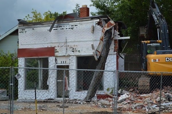 Damaged house with collapsed front wall beside a bulldozer and debris behind a chain-link fence