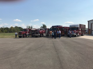 Red fire trucks and personnel gathered in a parking lot beside a fire station.