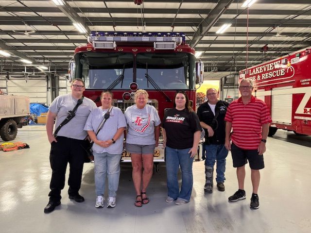 Six people pose in front of a red fire truck inside a fire station.