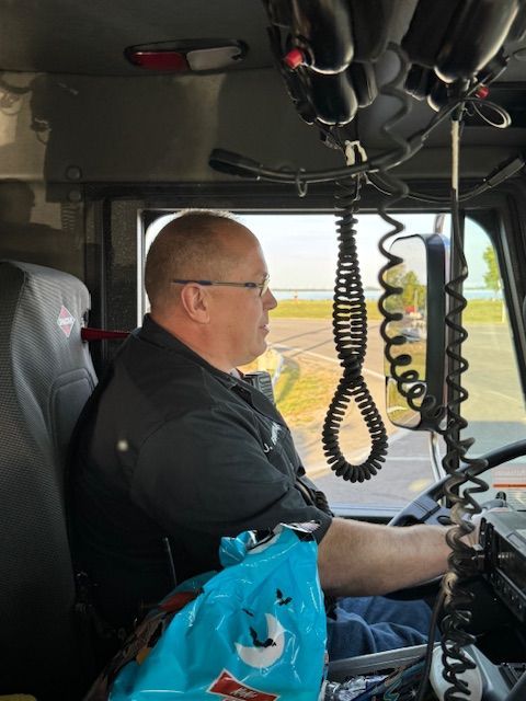 Person driving a truck cab, looking out the window, with coiled radio cords hanging overhead.