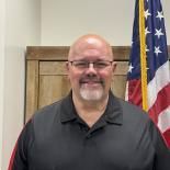 Smiling man in a dark polo shirt standing indoors beside a U.S. flag and wooden door.