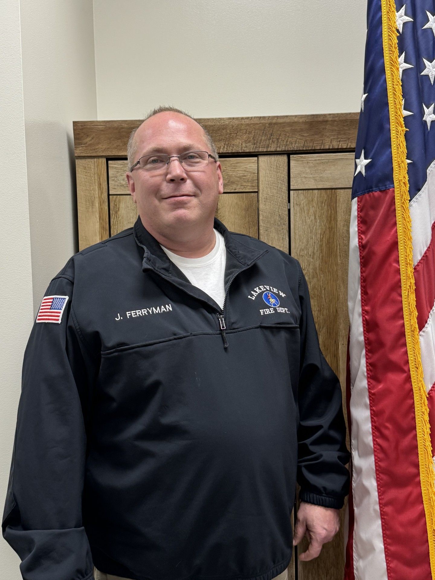 Man standing indoors beside an American flag, wearing a black jacket and smiling at the camera.