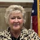 Portrait of a smiling woman in a leopard-print jacket, seated indoors beside a U.S. flag.