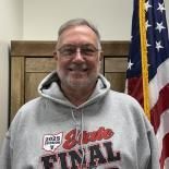 Smiling person in a gray hoodie stands indoors beside an American flag.