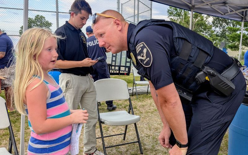 Police officer leaning down to speak with a young girl outdoors under a tent