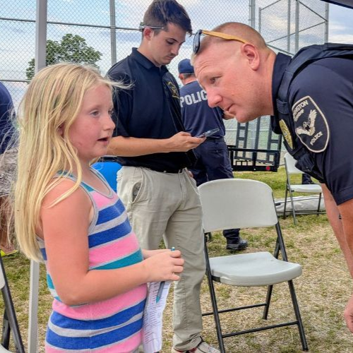 Police officer speaking with a young girl outdoors near a chain-link fence and folding chairs.