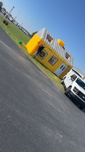 Yellow roadside kiosk on a tilted street beside a white vehicle, with a grassy field and blue sky behind.