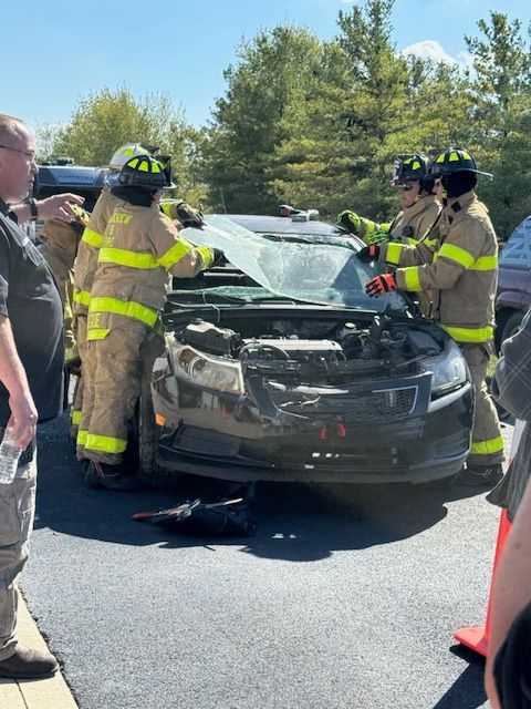 Emergency responders around a crashed black car with a crumpled hood on a road.