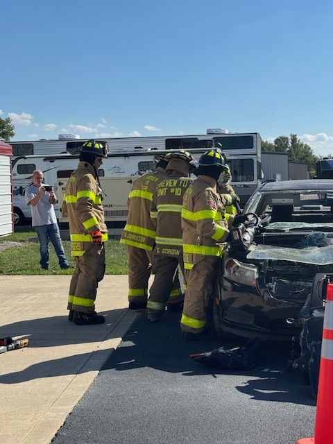 Firefighters in yellow gear at the rear of a burned car on a roadside, with smoke damage visible.