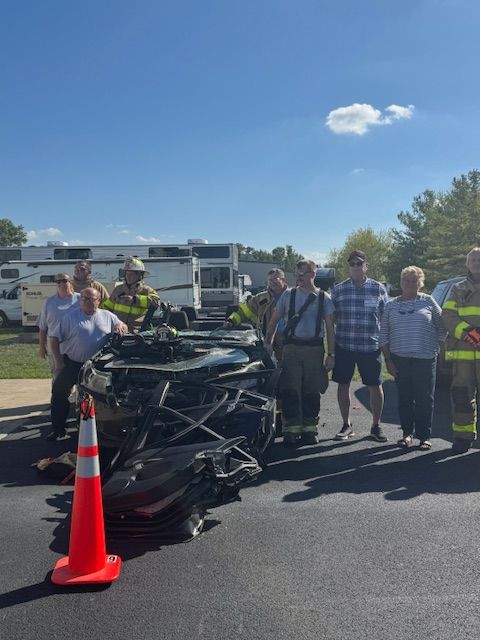 Crushed black car beside orange cone with group of people and emergency vehicles in a parking lot