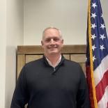 Smiling man in a black sweater standing indoors beside a U.S. flag