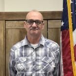 Man standing indoors in a plaid shirt and glasses, with a flag visible behind him.