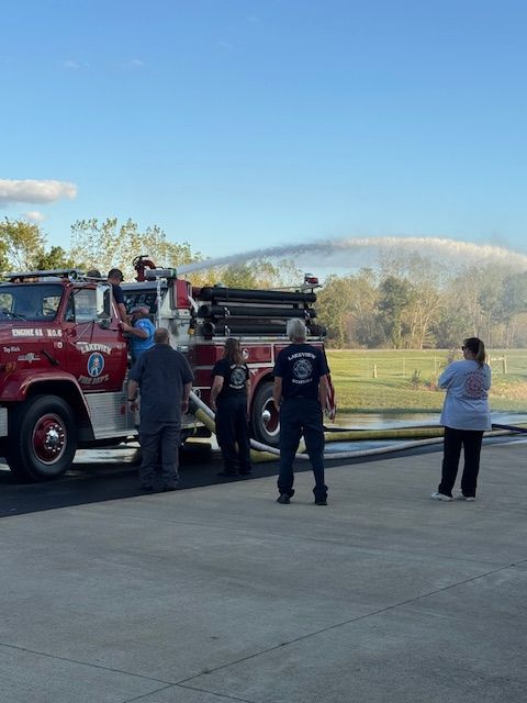 Firefighters beside a red fire truck at a roadside scene, with hoses laid out and smoke in the background