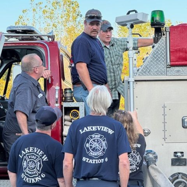 Lakeview firefighters gathered beside a red fire truck, with one man standing on the truck’s side step.