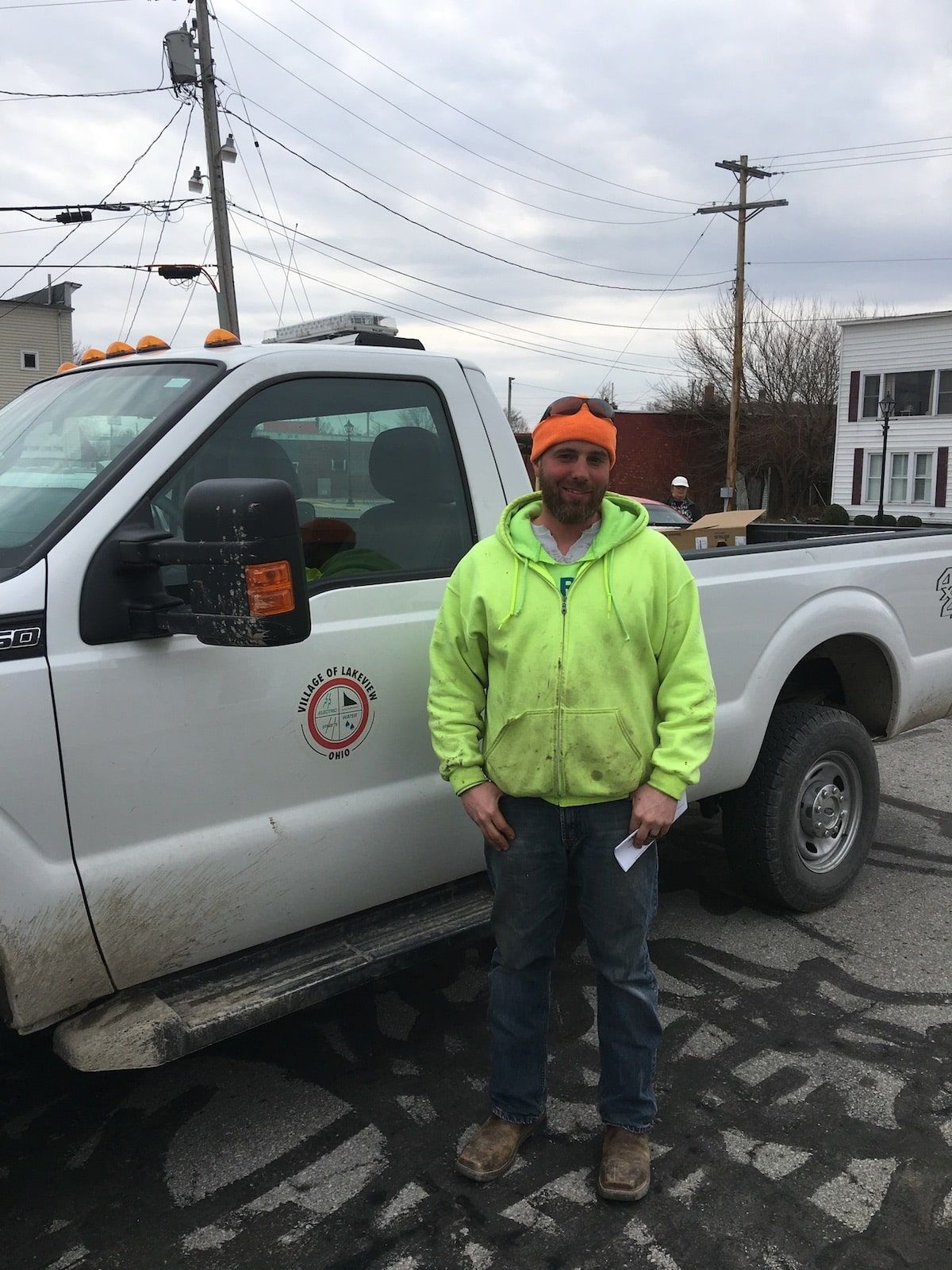 Worker in neon green jacket stands beside a silver pickup truck on a wet street.