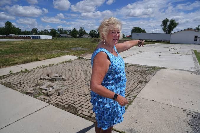 Woman in a blue dress points across a cracked paved lot toward low buildings under a cloudy sky
